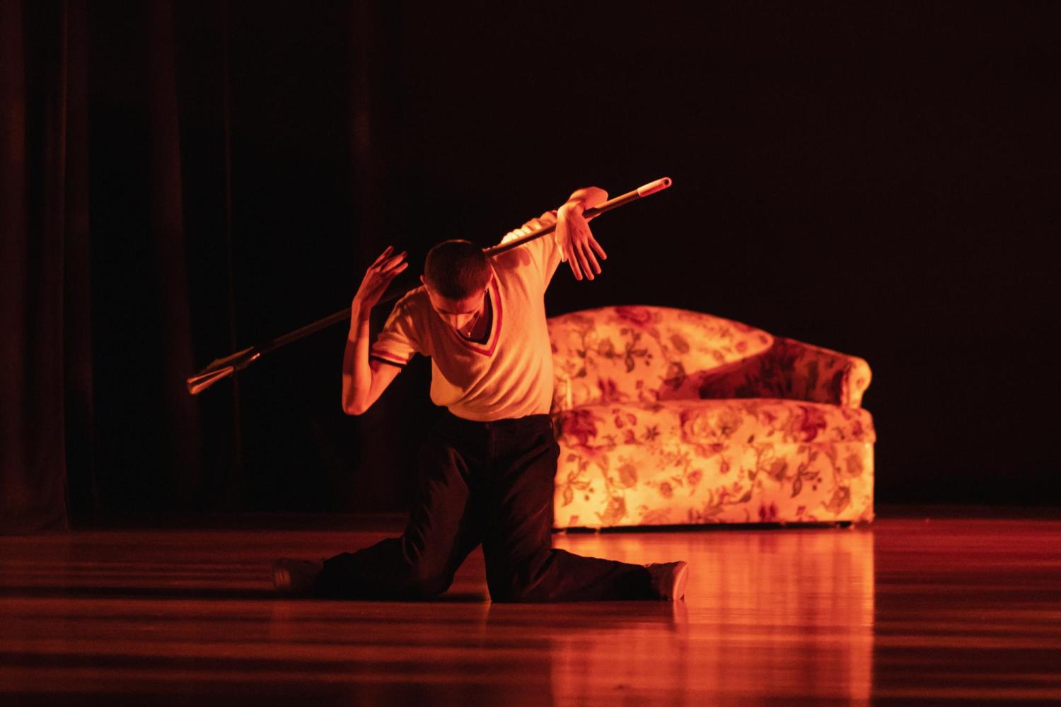 A photograph of James Solis Gutierrez performing dance. The photo is tinted red.He is posing dramatically with his knees to the ground, with his left arm outstreched behind him. In the background there is a flower-patterned couch.