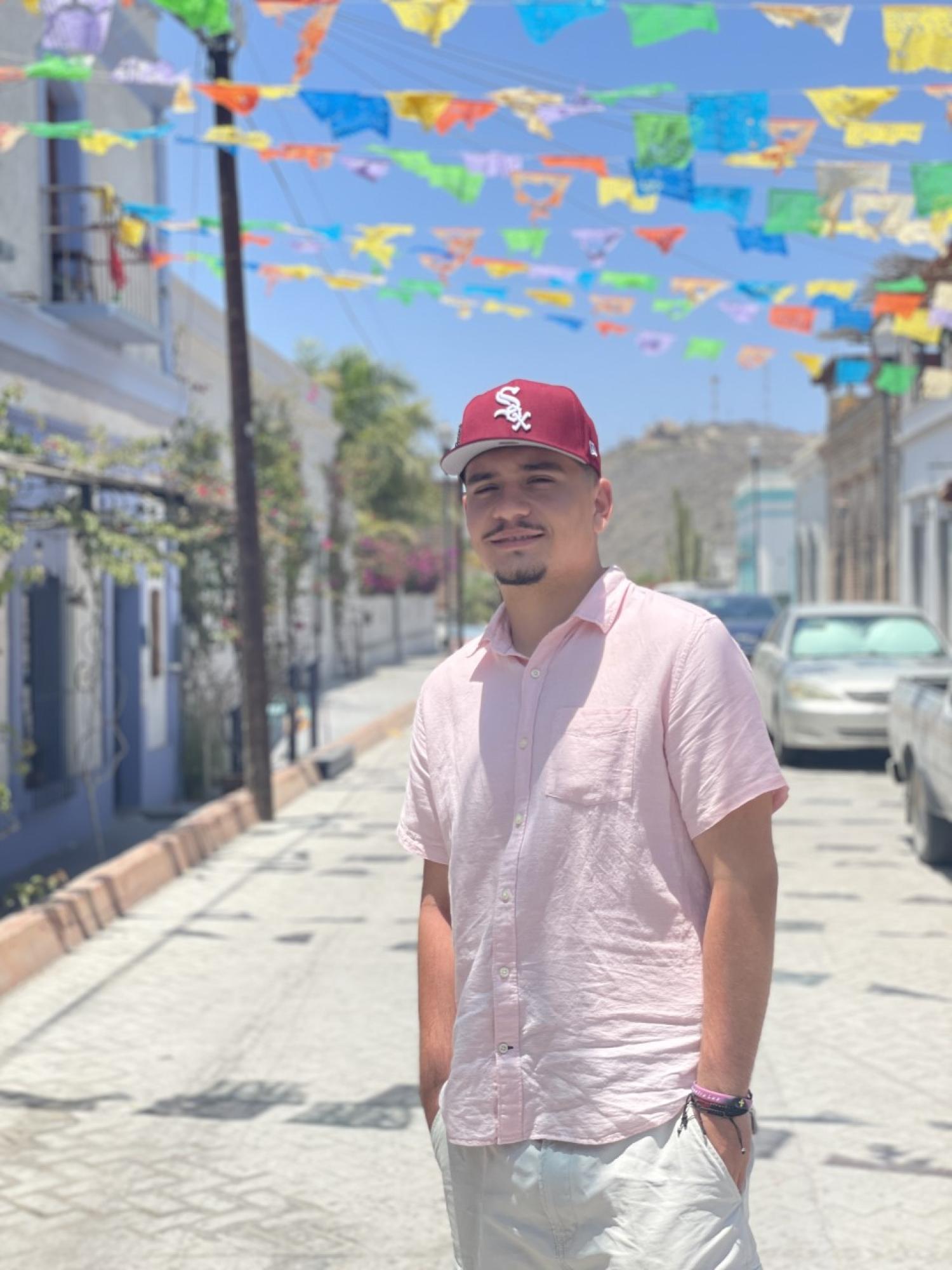 A photograph of Eduardo Torres. He is posing in a city street, wearing a light pink button-up shirt and a red Red Sox baseball cap.