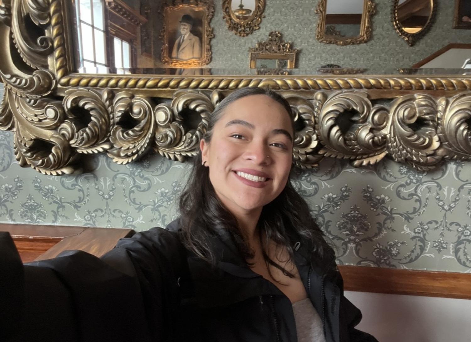 A photograph of Janeth Villalobos. She is wearing a black blazer, posing with her arm outstretched to hold the camera. Behind her is a greyish-green wall decorated in patterns, and a large bronze ornate mirror behind her.