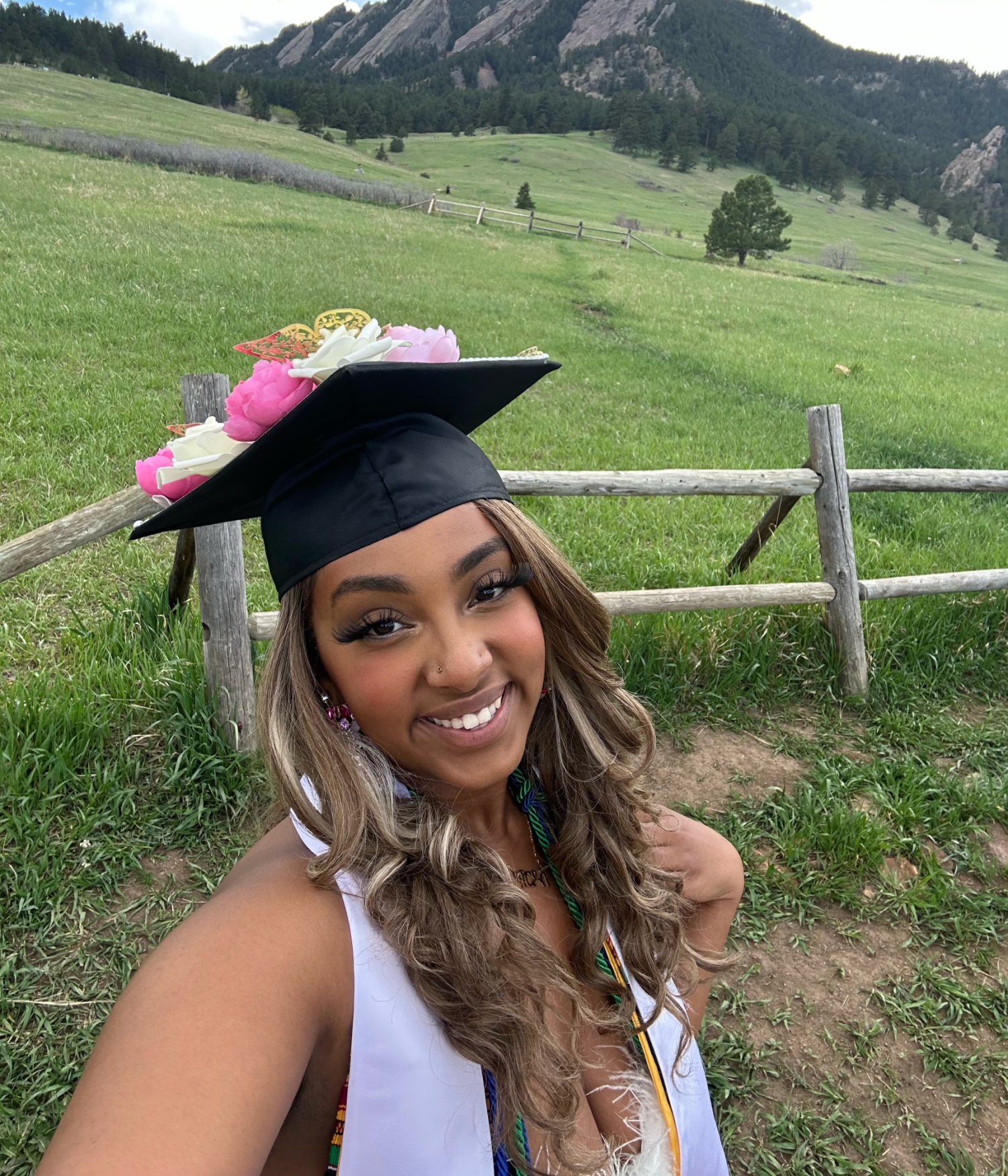 A photograph of Nardos Abayenh. She is wearing her graduate cap, decorated with flowers of varying shades of pink and white. She is posing in front of a large natural landscape, fields behind her and in the distance, tree covered hills and mountains.