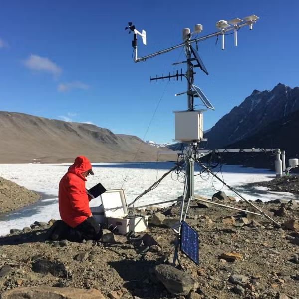 man kneeling down next to a weather sample tower with snow, mountains in background