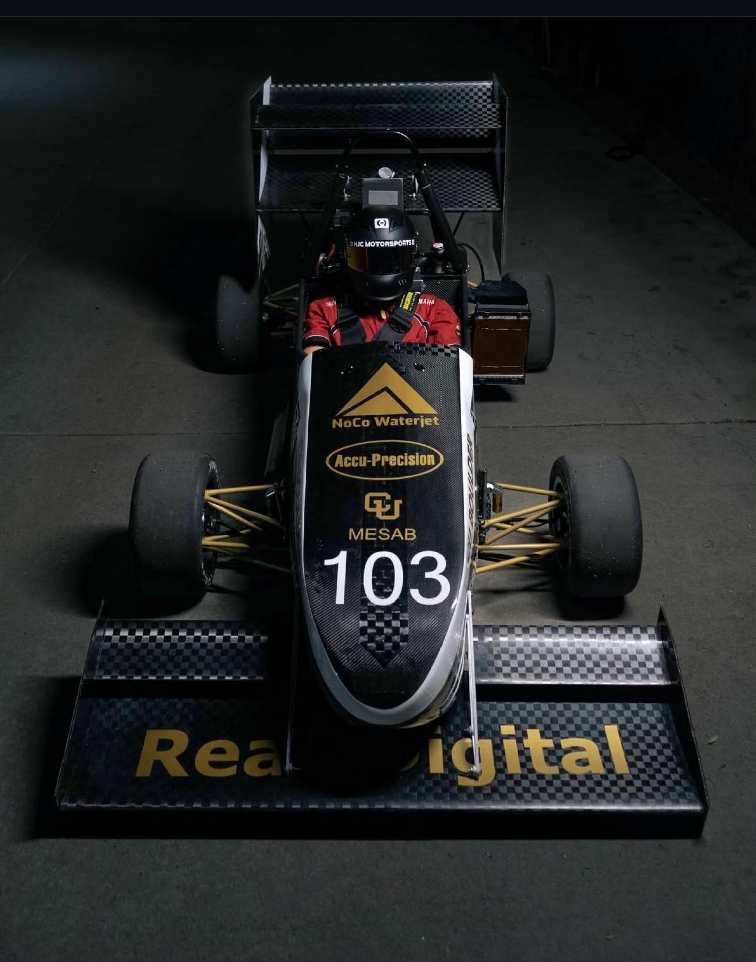 black FORMULA SAE vehicle with driver inside, dark background, CU Racing logo in front