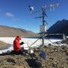man kneeling down next to a weather sample tower with snow, mountains in background