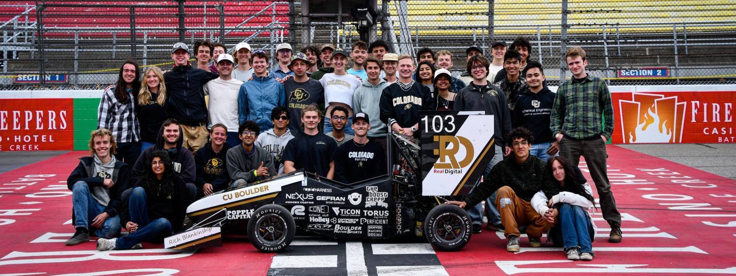 CU Boulder Racing team posing for group photo with Formula vehicle in front, at this year's Formula SAE competition in Michigan