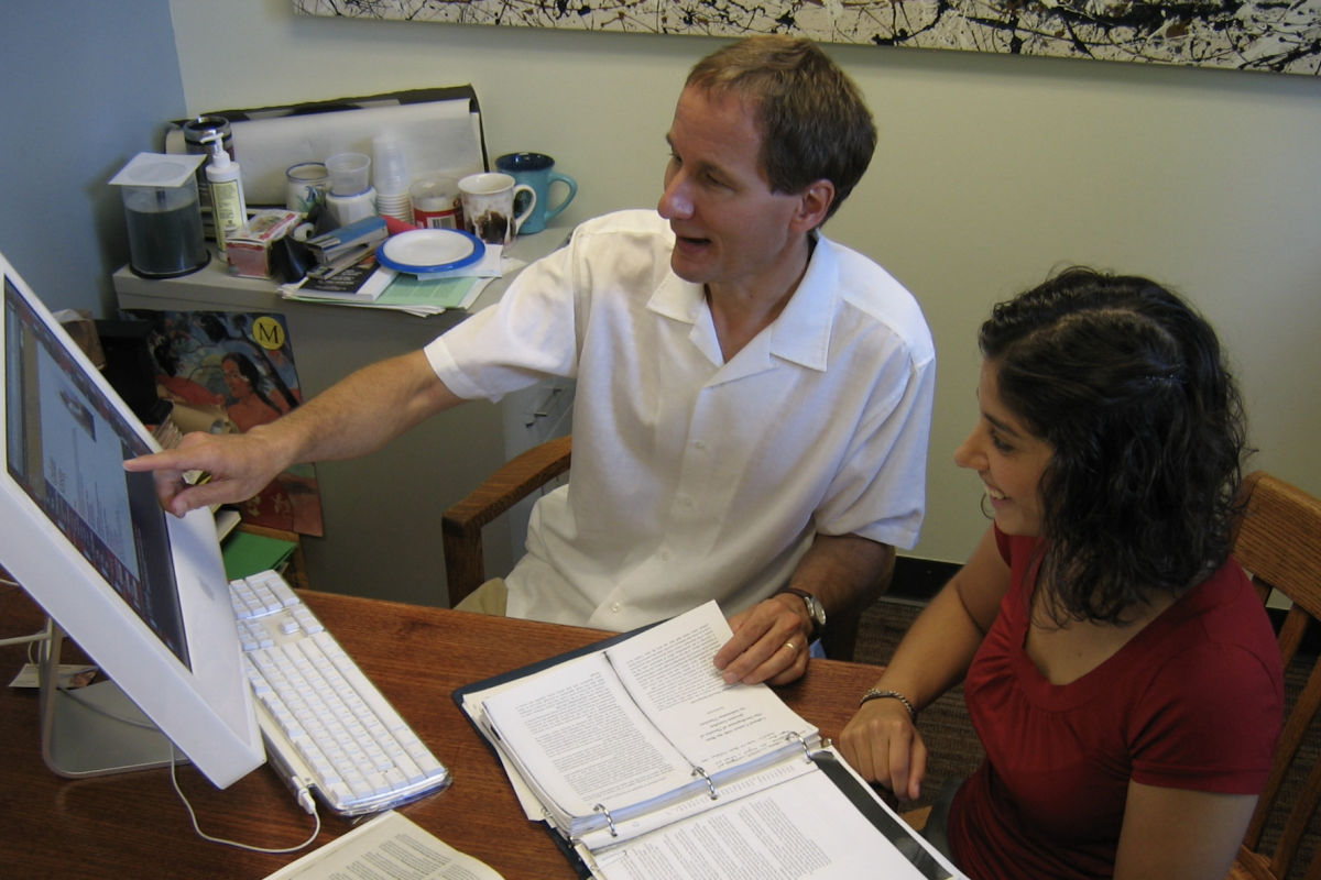 Man in white shirt teaching while sitting next to student looking at a computer screen.