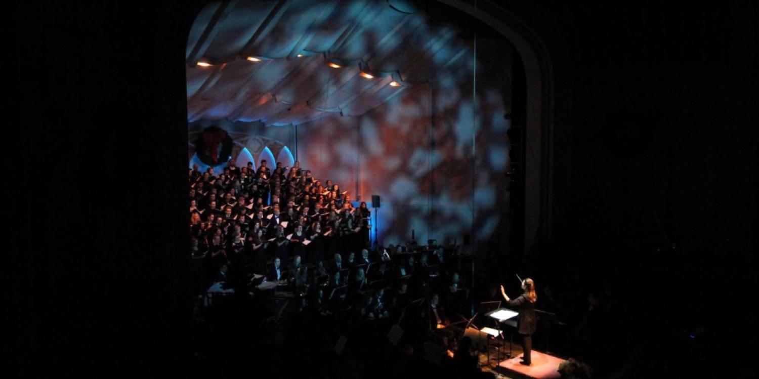 A conductor leads a choir and orchestra on a dimly lit stage with illuminated patterned projected on the ceiling