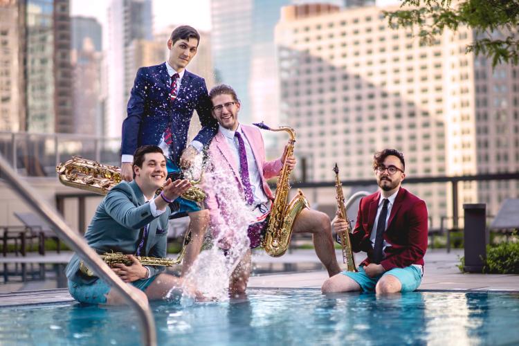 Four men in suits and shorts stand in a pool with saxophones. One splashes water as they pose against a cityscape background.