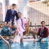 Four men in suits and shorts stand in a pool with saxophones. One splashes water as they pose against a cityscape background.