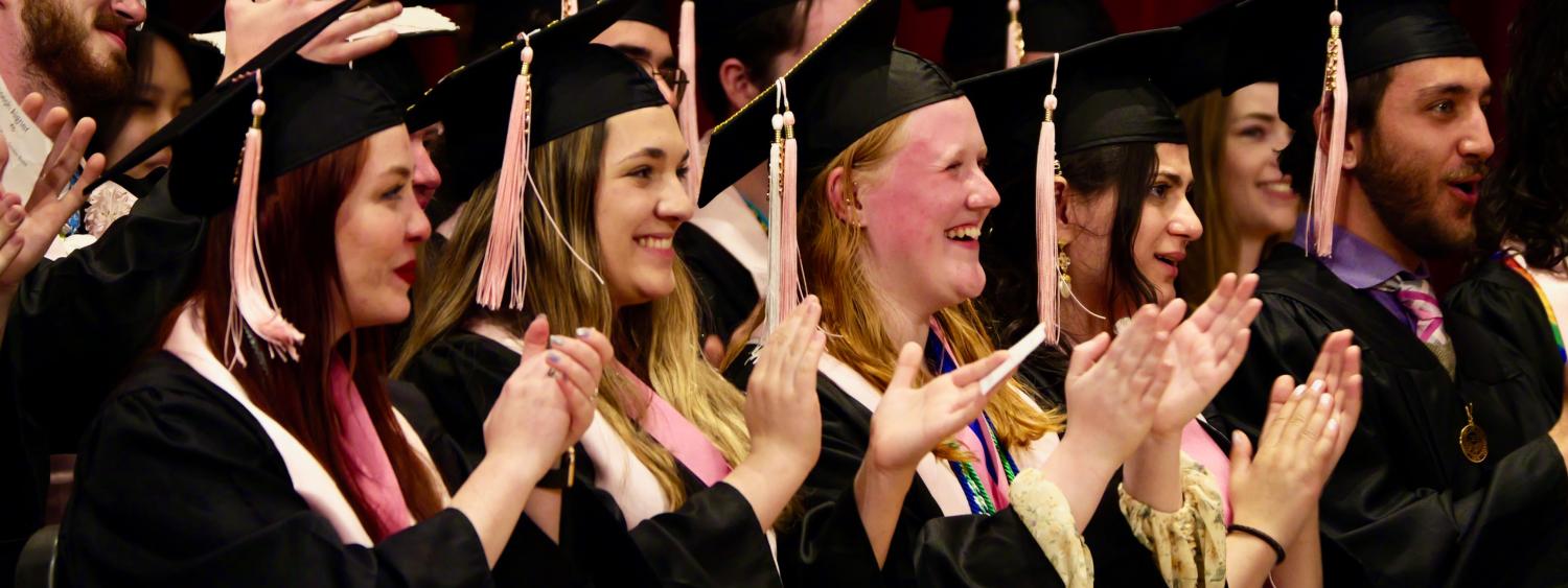 Students smiling at graduation ceremony in regalia while clapping