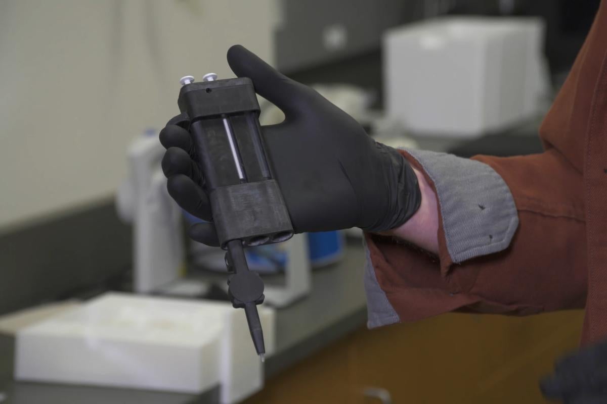 A small, handheld device about the size of a cell phone. A person wearing a black latex glove is holding the device.