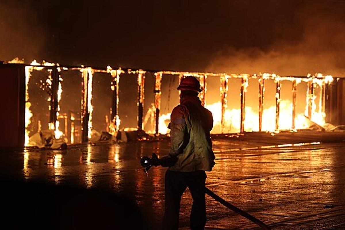 A firefighter stands in front of a building that has been reduced to its frame while flames continue to burn.