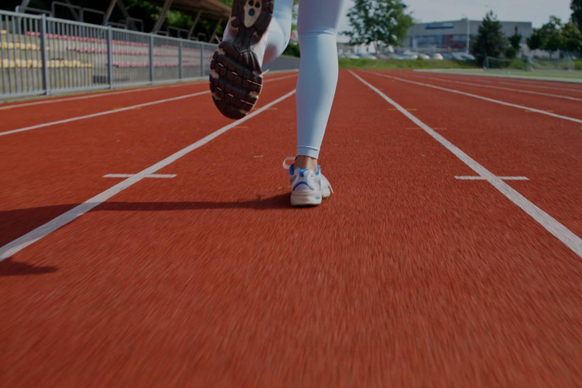 A person running on an outdoor track. The track is red and the person running is wearing blue shoes and leggings. The image is cropped and does not show the person above their knees.