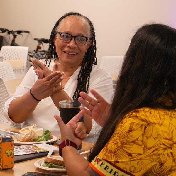 A female faculty member sits at a table across from another faculty member whose back is to the camera. Both are engaged in lively conversation