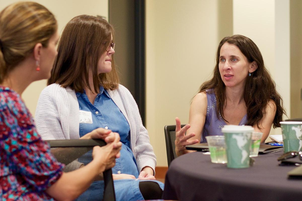 A woman sit at a table and chats with two other women across from her