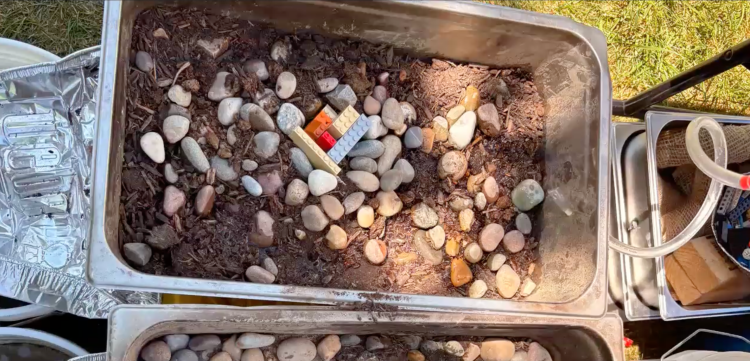A metal bin with strategically placed rocks and a small lego house to avoid damage from a simulated flood