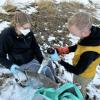Outside in the snow, a female researcher holds a bottle while a male researcher pours a surface water sample in it.