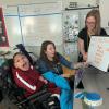 A teacher holds up a whiteboard with letters written out for it to an individual in a wheelchair who is smiling at the camera