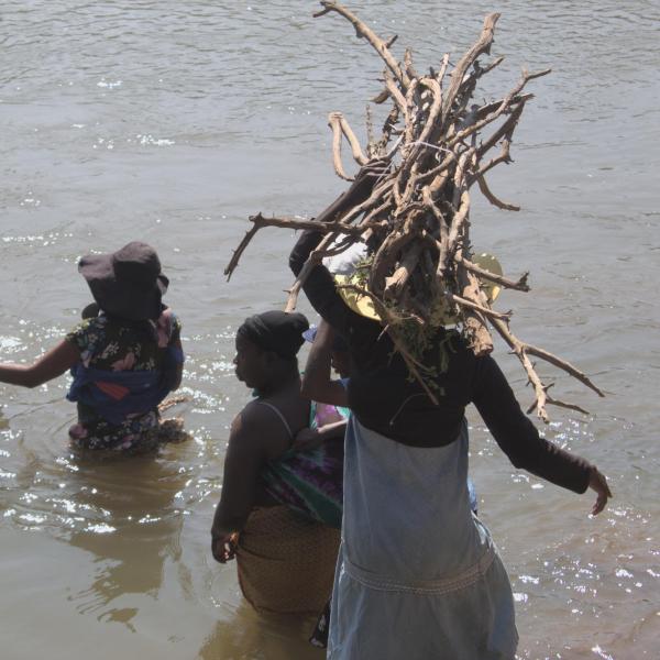 Community members carrying sticks and other items across the river, with the water coming up to their waist