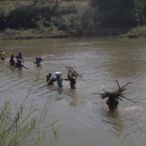 Another view of Community members carrying sticks and other items across the river, with the water coming up to their waist