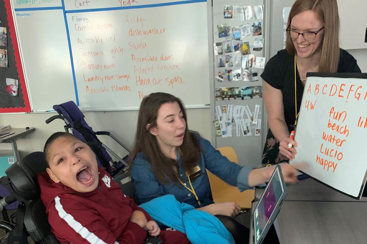 A teacher holds up a whiteboard with letters written out for it to an individual in a wheelchair who is smiling at the camera