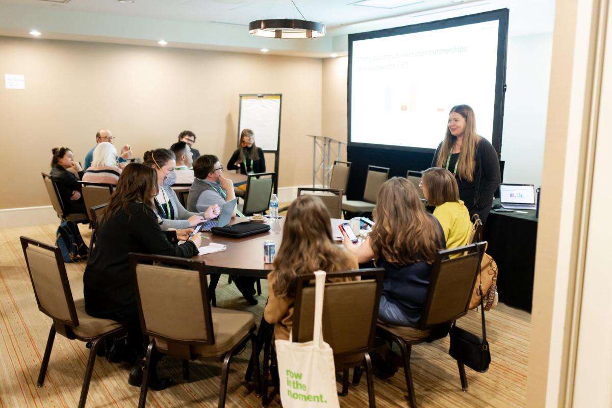 A woman stands in front of a screen, lecturing to a small audience in a conference room of round tables.