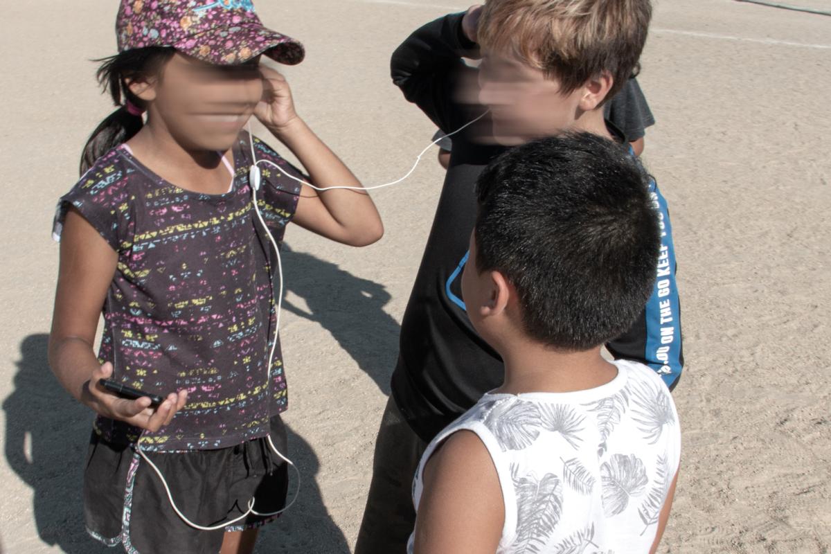 three children stand in a circle around each other. two of the children are sharing a pair of wired earbuds
