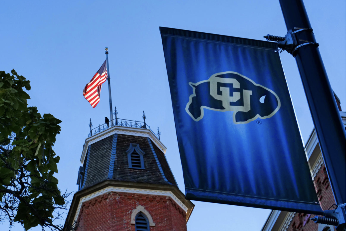 A U.S. flag flies near a red brick tower with crenellations. A banner featuring the black, gold, and white CU Boulder buffalo logo hangs from a pole in the foreground.