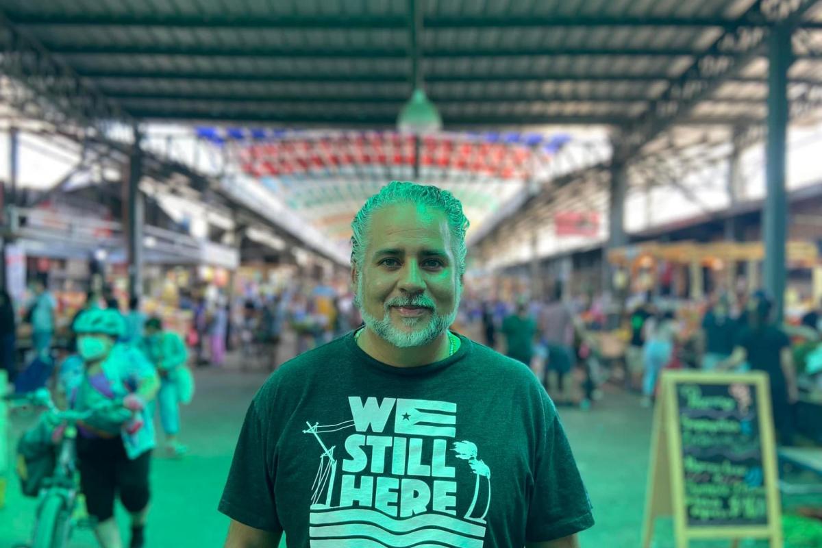 Khalil stands in front of a crowded market wearing a shirt that reads "We Still Here"
