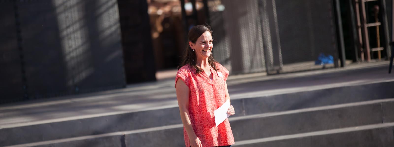 Amanda Giguere stands at the bottom of some steps outside somewhere. She is smiling and holding a piece of paper (mystery)
