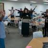 A female college student stands at the front of a room with her arms raised above her head. All other attendees i the room do the same.