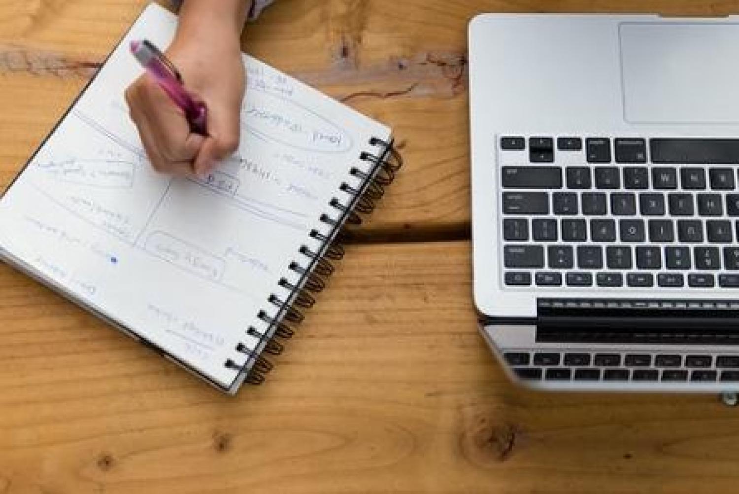 person's hand writing in planner next to laptop 