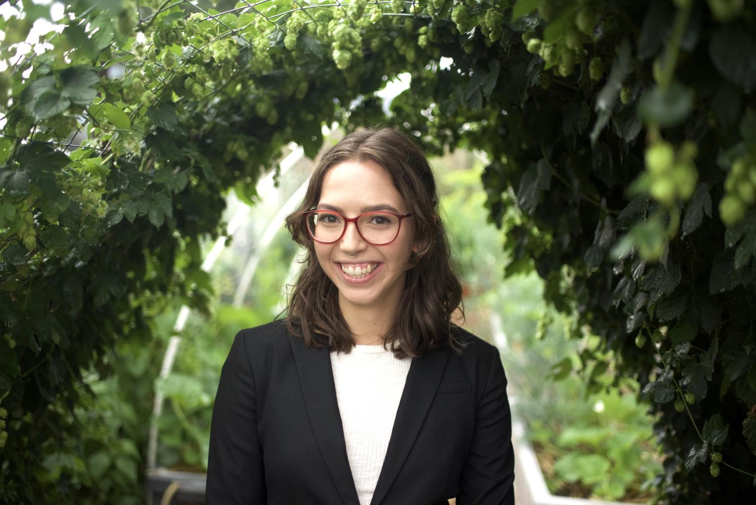 Image of Victoria - woman with shoulder length brown hair, glasses, wearing a black jacket and white shirt, and surrounded by a green leafy background