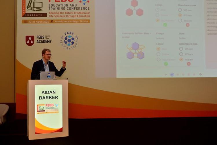 Aidan speaking on a stage at SciCon infront of a scrfeen presentation