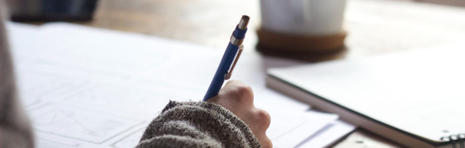 A person writing in a journal with a mug and a pad of paper next to them