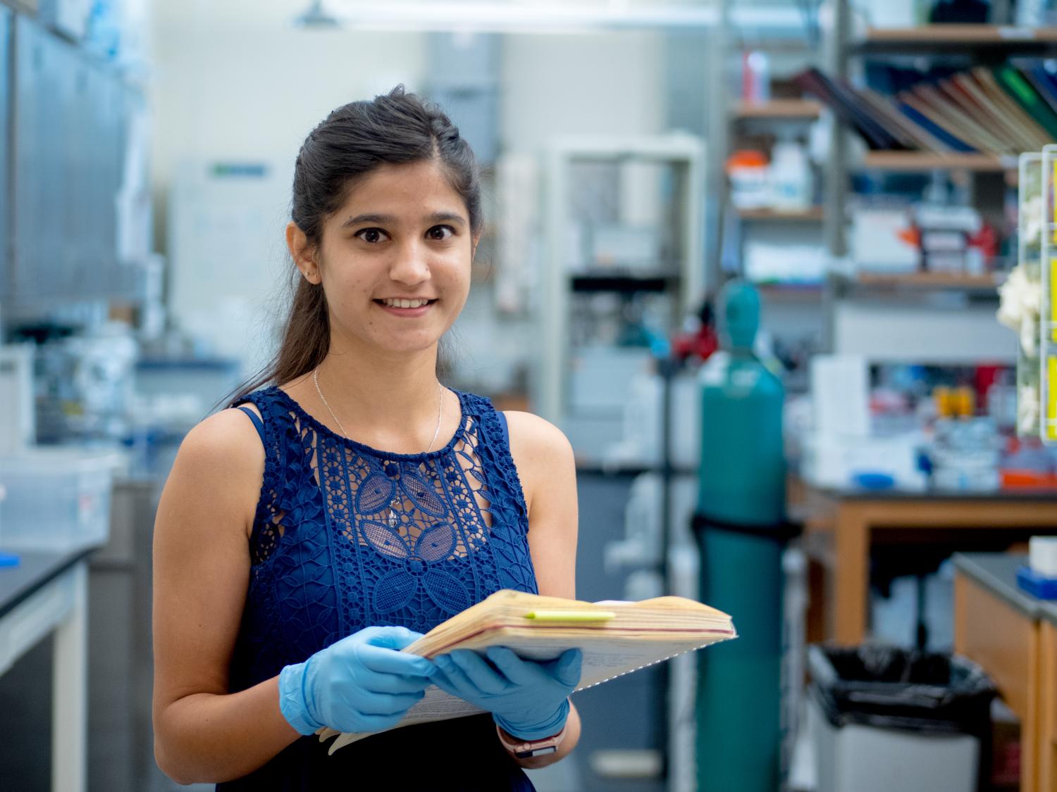 Student holds files of their cell biology research findings, smiling at a camera while standing in an elaborate lab setting.