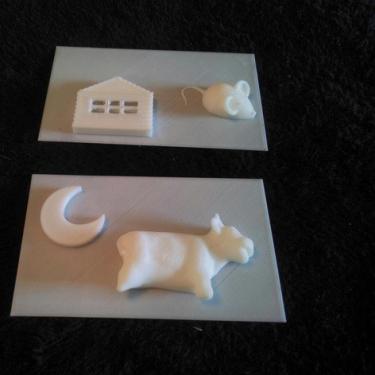 Two 3D printed pages showing relief of a cow with a moon; and a mouse with a house.