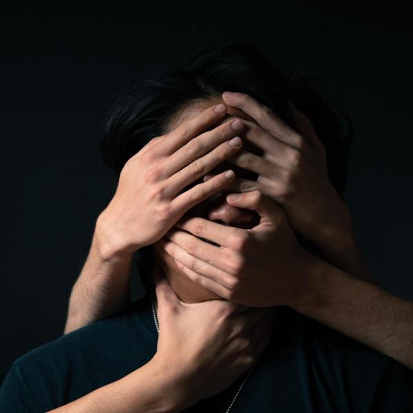 Headshot of man with long black hair with four hands grasping his face and neck with black background