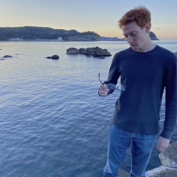 Cole Zucker - A person contemplates by the sea during sunset, holding sunglasses.
