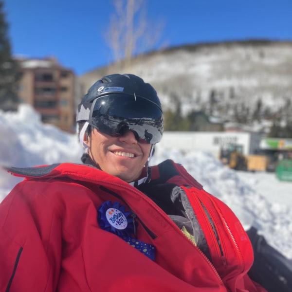 Michael McDermott - A smiling person wearing a ski helmet and goggles, with a snowy mountain backdrop, dressed in red ski gear