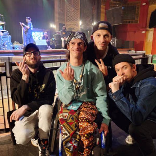 Sol Fuentes - A group of four individuals posing in front of a concert stage, giving peace signs and smiling