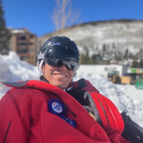 Michael McDermott - A smiling person wearing a ski helmet and goggles, with a snowy mountain backdrop, dressed in red ski gear