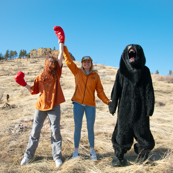 Sidney Yarnall - Two individuals and one person in a bear costume stand hand-in-hand, with the individuals raising one arm each, in a grassy field with a rocky outcrop in the background