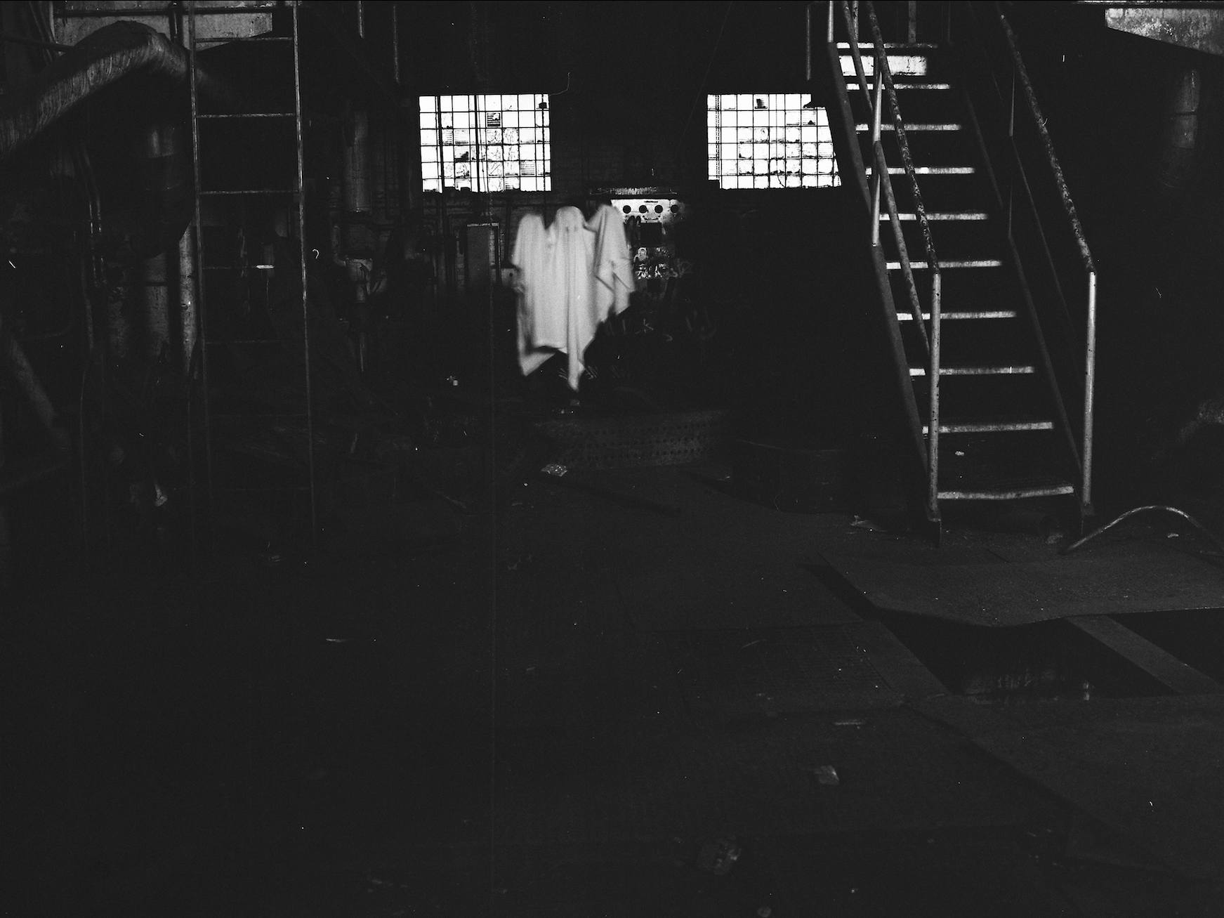 The image shows a dimly lit industrial interior with metal staircases, pipes, and machinery scattered in the darkness. Two barred windows at the back provide faint light, framing a ghostly figure draped in a white sheet standing in the shadows. The floor is cluttered with debris, while the rusted equipment and overall darkness enhance the eerie, abandoned atmosphere.