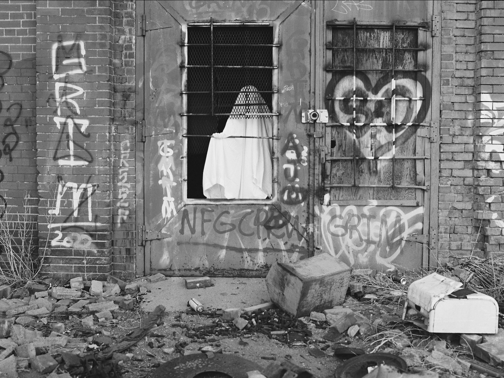 The image shows a dimly lit industrial interior with metal staircases, pipes, and machinery scattered in the darkness. Two barred windows at the back provide faint light, framing a ghostly figure draped in a white sheet standing in the shadows. The floor is cluttered with debris, while the rusted equipment and overall darkness enhance the eerie, abandoned atmosphere.