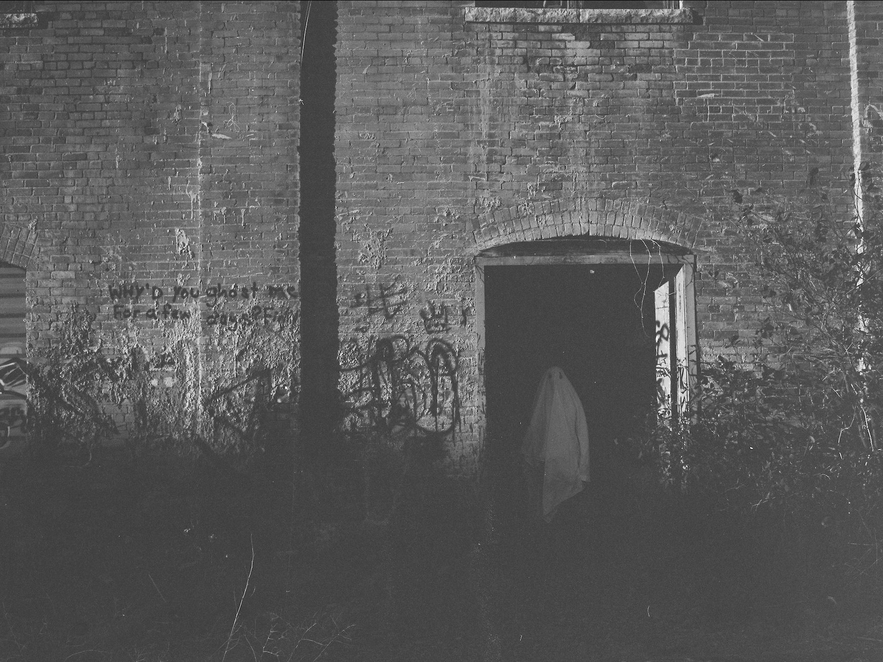 The image shows a dimly lit industrial interior with metal staircases, pipes, and machinery scattered in the darkness. Two barred windows at the back provide faint light, framing a ghostly figure draped in a white sheet standing in the shadows. The floor is cluttered with debris, while the rusted equipment and overall darkness enhance the eerie, abandoned atmosphere.