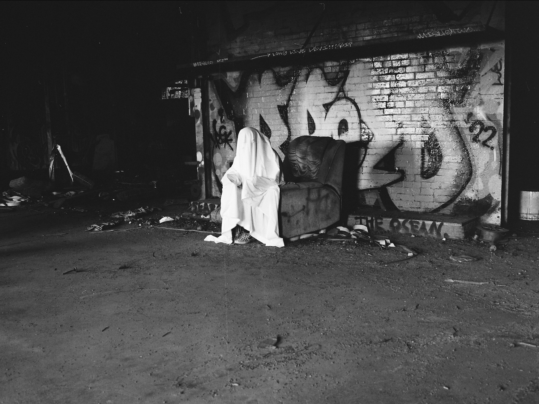 The image shows a dimly lit industrial interior with metal staircases, pipes, and machinery scattered in the darkness. Two barred windows at the back provide faint light, framing a ghostly figure draped in a white sheet standing in the shadows. The floor is cluttered with debris, while the rusted equipment and overall darkness enhance the eerie, abandoned atmosphere.