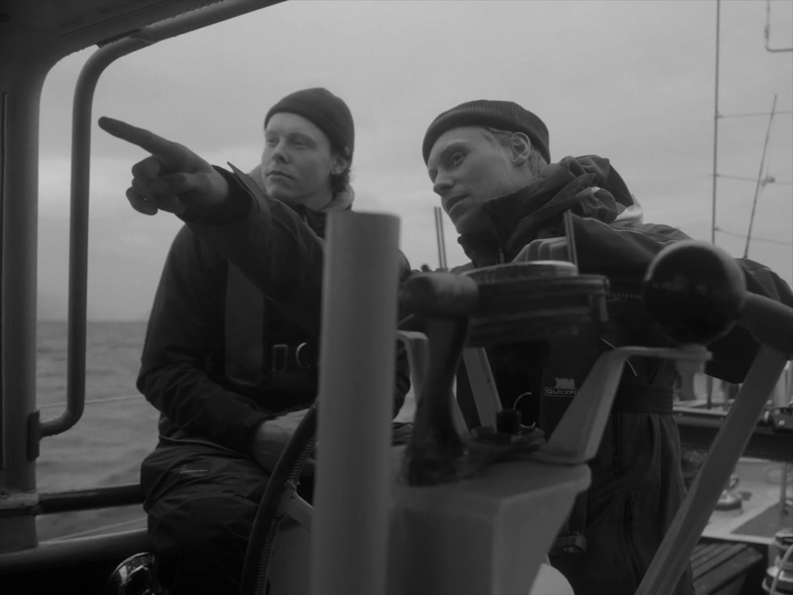 B&W two sailors behind wheel of sailboat, one pointing