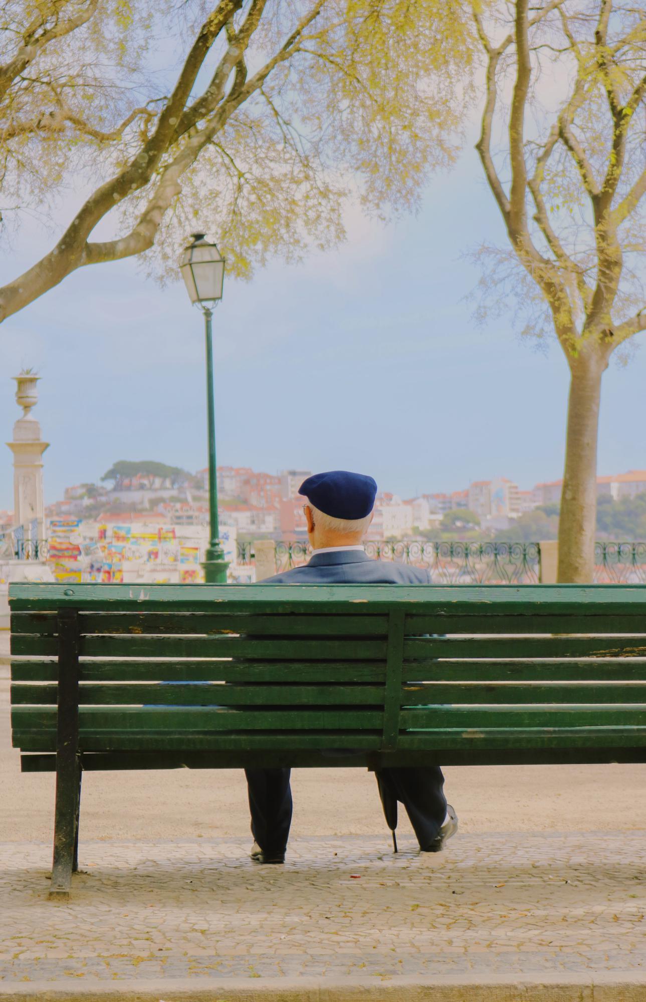 An older gentleman sits at a green park bench, clutching an umbrella, and admiring a sunny, scenic view of Lisbon.