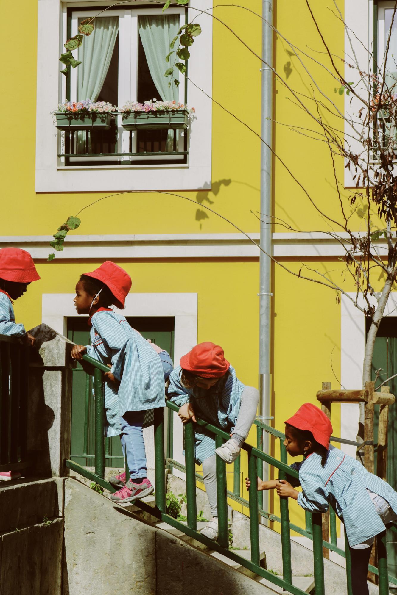 school children, in matching uniforms, play on a stairwell in front of a vibrant yellow building