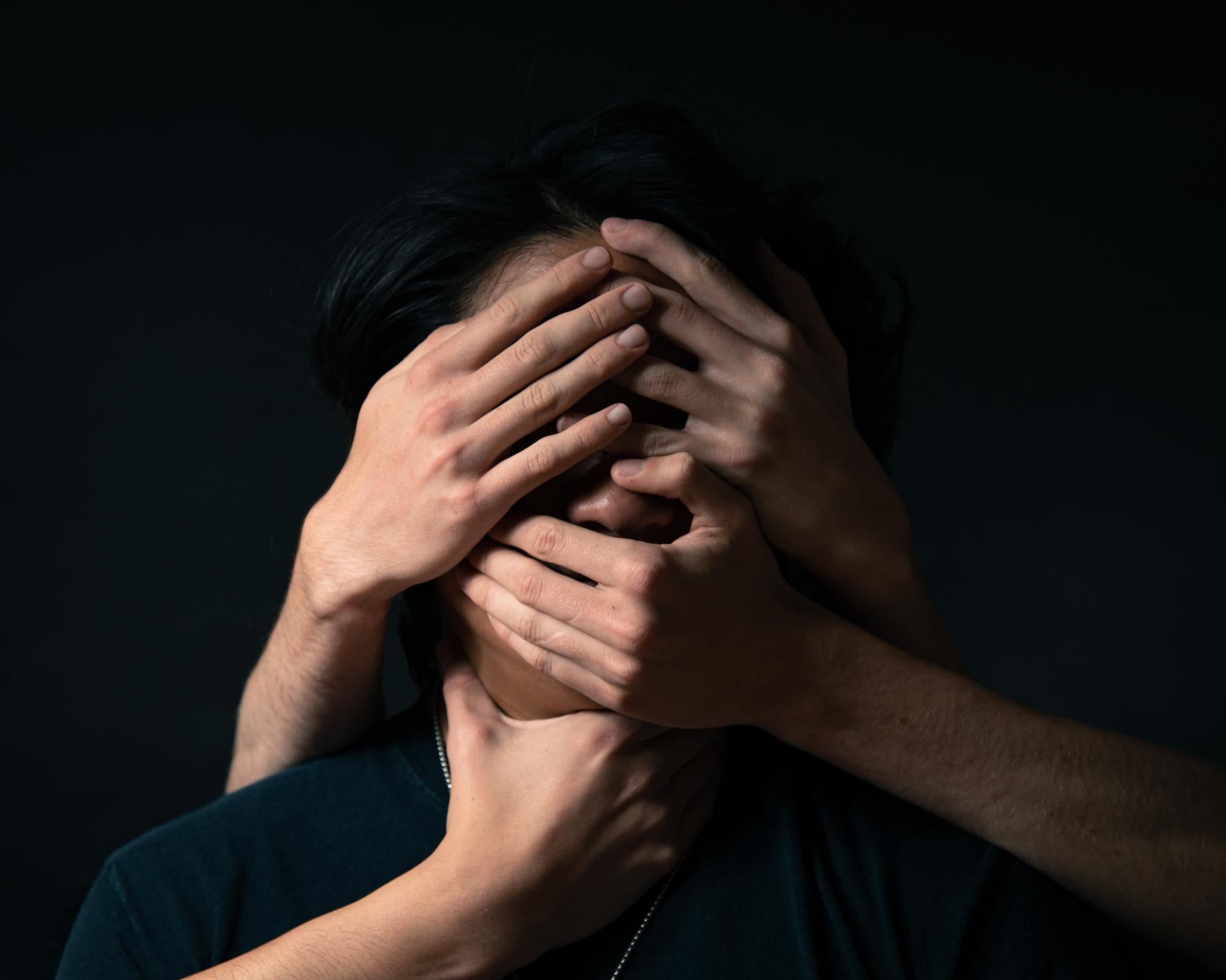 Headshot of man with long black hair with four hands grasping his face and neck with black background